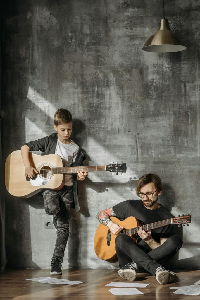 A father and son share a musical moment, playing guitars indoors with warm lighting.