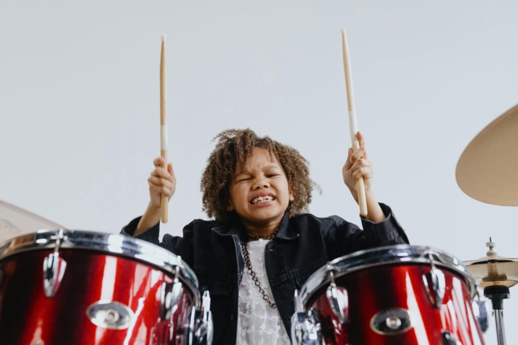 Young child joyfully playing drums with energetic expression and drumsticks raised high.